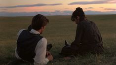 two people sitting on the ground in an open field at sunset, one holding a cell phone