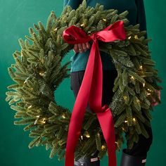 a person holding a christmas wreath with red ribbon and lights on the front, against a green background