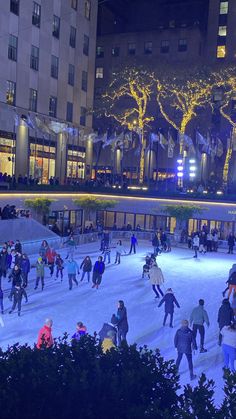 people skating on an ice rink at night in front of tall buildings with christmas lights