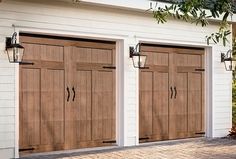 three wooden garage doors in front of a white house with brick flooring and trees