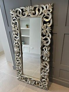 an ornate silver framed mirror on the floor in front of a gray wall with shelves