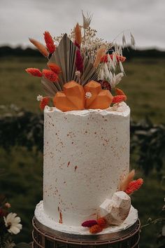 a white cake with orange flowers and feathers on top is sitting in the middle of a field