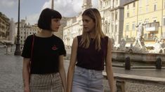 two young women standing in front of a fountain on a cobblestone street with buildings in the background