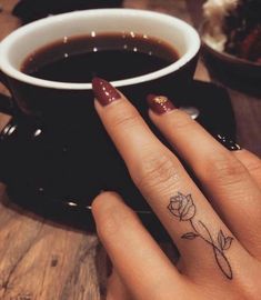 a woman's hand with a small rose tattoo on her ring finger next to a cup of coffee