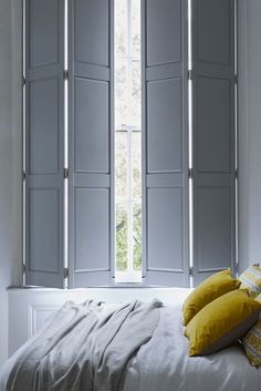 a white bed sitting under two windows with shutters on each side and yellow pillows