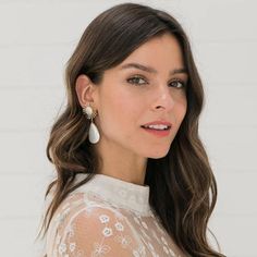 a woman with long brown hair wearing earrings and a white top is looking at the camera