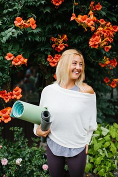 a woman is holding a yoga mat and smiling at the camera while standing in front of flowers