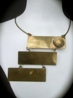 1970's Avant-garde brass metal articulated tile choker necklaceThe unique artisan choker necklace is designed with three large scale brass metal rectangular articulated tilesThe trio of brass metal tiles are suspended from a rigidwire band that wraps around the neck. One of the brassmetal rectangular tiles is adorned with a milky smooth glass cabochonThe edgy mod handmade artisan choker makesa very chic one of a kind accessory Measurements The rigid brass metal wire band interior circumferenceth Brutalist Brass Jewelry In Bronze, Antique Rectangular Brass Jewelry, Brutalist Bronze Brass Jewelry, Vintage Brass Rectangular Jewelry, Vintage Rectangular Brass Jewelry, Adjustable Rectangular Bronze Jewelry, Bohemian Brass Rectangular Jewelry, Vintage Square Metal Jewelry, Unique Bronze Rectangular Jewelry