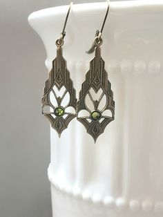 These modern Deco earrings are a great example of classic 1920s style. They feature ornate Art Deco style brass pendants. The pendants have a tiny olivine green crystal at the center These elegant earrings measure 2 inches long from the top of the ear wires to the bottom of the pendants. I love the simplicity of these Art Deco inspired earrings; perfect for everyday or evening. ABOUT THIS COLLECTION What are the three style essentials Jazz Age flappers like Clara Bow, Louise Brooks, and Zelda Fi Flapper Jewelry, Art Deco Jewelry 1920s, 1920s Earrings, Gold Art Deco Earrings, Gatsby Jewelry, Phoenix Jewelry, Clara Bow, Louise Brooks, Brass Pendants