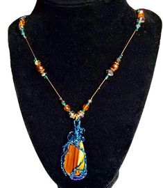 Embrace the grounding and protective energies of Tiger's Eye with this stunning handmade pendant. This unique piece features a polished Tiger's Eye stone, known for its beautiful bands of golden brown and vibrant hues. The stone is intricately wrapped in teal wire, adding an artistic and modern touch to the natural beauty of the gemstone. The pendant is complemented by a meticulously beaded necklace, showcasing a harmonious blend of amber, teal, and clear beads that catch the light beautifully. Each bead is carefully selected to enhance the overall aesthetic, making this necklace a perfect statement piece for any outfit. Tiger's Eye is believed to bring clarity, focus, and confidence to its wearer. Whether you're looking for a meaningful gift or a special addition to your jewelry collectio Bohemian Crystal Necklaces With Natural Stones In Copper, Bohemian Copper Crystal Necklaces With Natural Stones, Spiritual Hand Wrapped Orange Jewelry, Nature-inspired Brown Jewelry For Festivals, Hand-wrapped Jasper Pendant Necklace, Agate Wire Wrapped Jewelry For Meditation, Spiritual Jasper Necklaces With Wire Wrapped, Spiritual Jasper Necklaces Wire Wrapped, Hand Wrapped Jasper Pendant Necklace