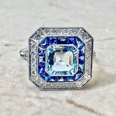 Breathtaking handcrafted platinum Art Deco style aquamarine, sapphire and diamond ring! The center stone is a lovely 1.05 carat aquamarine. It is surrounded by a row of fine French cut sapphires weighing 0.70 carat. The sapphires are haloed by 0.40 carat of round diamonds. Fine milgrain adds to the delicacy of the ring. Beautiful openwork filigree decorate the under gallery. The ring measures 13.97 x 13.63 mm from side to side. Stamped Pt900. Ring size 6.25-6.5 US / M-M 1/2 UK. > Resizing includ Blue Art Deco Diamond Ring With Accents, Blue Art Deco Diamond Ring With Center Stone, Art Deco Blue Diamond Ring With Accents, Art Deco Blue Diamond Ring With Center Stone, Art Deco Blue Sapphire Ring With Center Stone, Blue Platinum Art Deco Diamond Ring, Blue Diamond Art Deco Ring, Blue Art Deco Diamond Ring, Blue Art Deco Jewelry Gia Certified