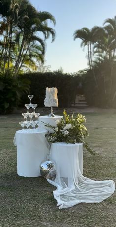 a table set up with white linens and silver vases on the grass in front of palm trees