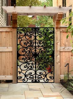 an iron gate in front of a wooden fence with plants on the side and trees behind it