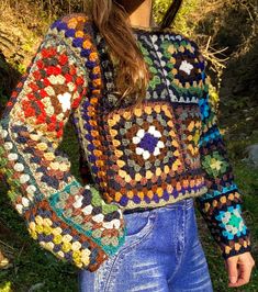 a woman wearing a multicolored crocheted sweater and jeans standing in the grass