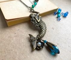 "This is a whimsical necklace with a large mermaid pendant. There is a teal faceted glass bead, a glass flower bead and a seashell dangling from the mermaid's tail. An unusual necklace, handcrafted by Selma Dreams in Finland. The necklace is made of antiqued brass, all parts are lead and nickel safe. The sturdy brass chain with the filigree adorned glass beads is 80cm/31\" long, although the length can also be altered to your needs. If you have any questions, please do ask.  The necklace will be Handmade Mermaid-shaped Ocean-inspired Necklaces, Bohemian Mermaid Necklace For Gifts, Bohemian Mermaid Necklace Gift, Ariel Necklace, Whimsical Mermaid, Whimsical Necklace, Large Mermaid, Teal Glass, Handmade Mermaid