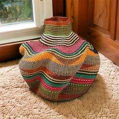 a multicolored basket sitting on the floor in front of a window, next to a rug