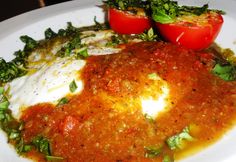 a white plate topped with meat covered in sauce and veggies next to tomatoes