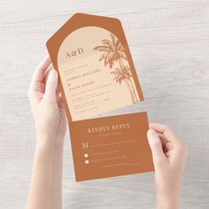 two hands holding up a brown and white wedding card with palm trees in the background