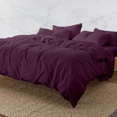 a bed with purple sheets and pillows on top of a brown rug in front of a white wall