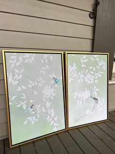 two framed paintings sitting on top of a wooden floor next to a wall with white flowers