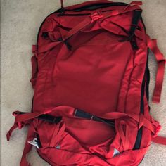 a red backpack laying on the floor with straps around it's sides and zippers at the bottom