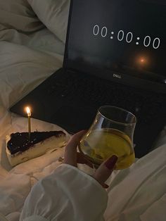 a person holding a lit candle next to a piece of cake on a bed with a laptop in the background