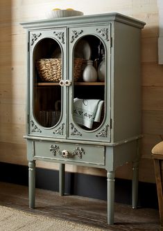 a green cabinet with two glass doors and some baskets on top of it, in front of a wooden wall