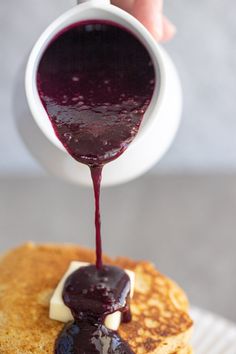 someone pouring blueberry sauce on top of a stack of pancakes with butter and syrup