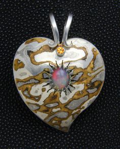 This heart pendant is made from Mokume Gane, a process of metal fabrication that dates back to the age of the Samurai. It involves fusing two or more alloys that are then carved, twisted, sanded, or otherwise manipulated to produce a pattern similar to wood grain. In this case, the metals I used were sterling silver, brass, and a Japanese alloy called Shibuichi. In the center, as though bursting from a volcano, is an African Welo Opal. The accent stone at the top, beneath the scrolls in the bail, is a faceted citrine. The pendant measures 2 inches from the tip to the top of the bail. Artisan Engraved Heart Jewelry, Artisan Engraved Heart-shaped Jewelry, Unique Hand Forged Necklace With Heart Pendant, Unique Hand Forged Heart Pendant, Unique Hand Forged Heart Pendant Jewelry, Unique Hand Forged Heart-shaped Jewelry, Artisan Heart-shaped Jewelry For Anniversary, Silver Hand Forged Heart Necklace, Hand Forged Silver Heart Necklace