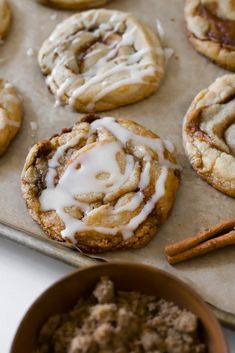 cinnamon roll cookies with icing and cinnamon sticks on a baking sheet next to bowl of cinnamon rolls