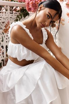 a woman in a white dress leaning against a wall with pink flowers behind her,