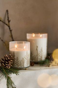 two white candles sitting on top of a table next to pine cones and evergreen branches