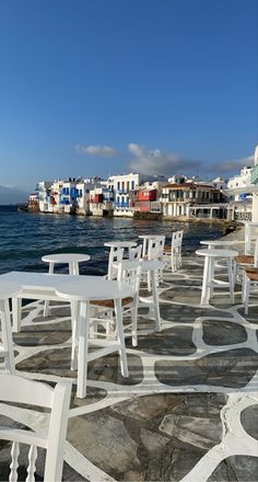 white tables and chairs sitting on top of a stone walkway next to the ocean with buildings in the background