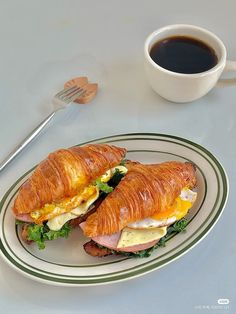 there is a croissant sandwich on the plate next to a cup of coffee