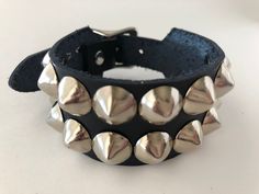 "Classic studded punk bracelet with 2 rows of 1/2\" cone studs. Fastens with a buckle and is adjustable. All hardware is nickel plated. Studs on black bracelets are nickel plated brass, studs on colored bracelets are nickel plated steel (old stock) Smaller bracelets have fewer studs per row. Made with vegetable tan leather. Note: red bracelet is kind of pink-ish. Bracelet is 1.25\" wide. Ask about custom sizing and color options!" Adjustable Punk Cuff Bracelet With Rivets, Punk Adjustable Cuff Bracelet With Rivets, Punk Style Adjustable Cuff Bracelet With Rivets, Punk Black Bracelet With Silver Studs, Trendy Adjustable Spiked Bracelets, Silver Punk Bracelets With Spikes, Silver Punk Bracelets With Studs, Punk Leather Bracelet With Studs For Festivals, Punk Style Silver Bracelet With Studs