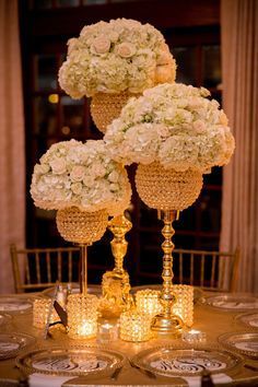 three tall vases filled with white flowers on top of a gold table cloth covered table