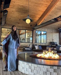 a woman standing next to a fire pit in a living room with wooden floors and walls