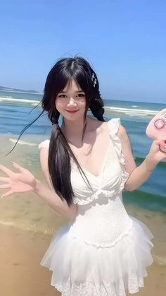 a woman in white dress standing on beach holding up a pink object with her hands