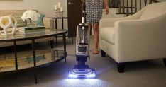 a woman standing in a living room with a blue and white vacuum cleaner on the floor