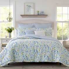 a white bed with blue and yellow flowers on it in a room next to two windows