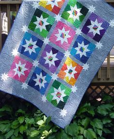 a colorful quilt hanging from a wooden fence next to green plants and bushes on a sunny day