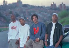 three men standing next to each other in front of a car with the city behind them