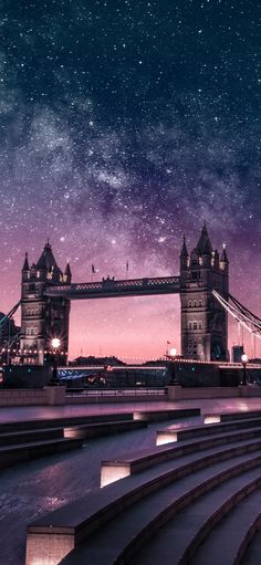 the tower bridge is lit up at night with stars in the sky above it and water below