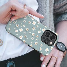 a woman holding an iphone case with white daisies on it, in front of her is a watch