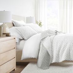 a bed with white sheets and pillows in a bedroom next to a dresser, lamp and window