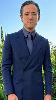 a man wearing a suit and tie standing in front of some bushes with his hands in his pockets