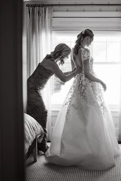 two women in dresses are standing by a window