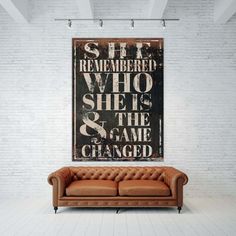 a brown couch sitting in front of a poster on a white brick wall next to a wooden floor