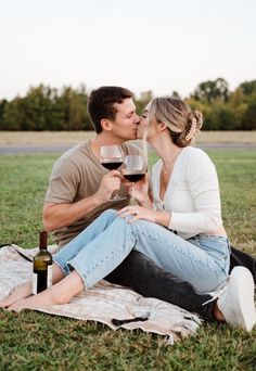 a man and woman sitting on the grass kissing while holding wine glasses