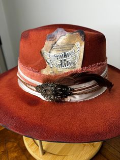 "The perfect burnt orange (rust) hat has been hand painted, and distressed. It's a smaller fedora and has a pink velvet band, wrapped in lace and gold threading. A vintage iron pin is attached. Tied with pink twine is a red feather and brown duck tail feathers.   SIZE: Fedora Hat is OSFA Wired brim measuring 3 3/4\" and a crown height of 4 3/4\". The Fedora Hat has a adjustable band with drawstring which allows for a perfect fit. Materials: This hat is made from a blend of 65% cotton and 35% pol Vintage Handmade Hat Band For Western-themed Events, Vintage Handmade Hats For Western-themed Events, Vintage Handmade Hat For Western-themed Events, Vintage Handmade Hats For Country Events, Handmade Vintage Hats For Western-themed Events, Vintage Distressed Hats For Rodeo, Vintage Handmade Adjustable Felt Hat, Handmade Adjustable Vintage Felt Hat, Handmade Adjustable Vintage Top Hat